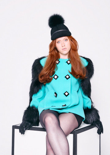 Person wearing a turquoise sweater with black diamond patterns, black fur vest, and black hat with pom-pom on a white background.